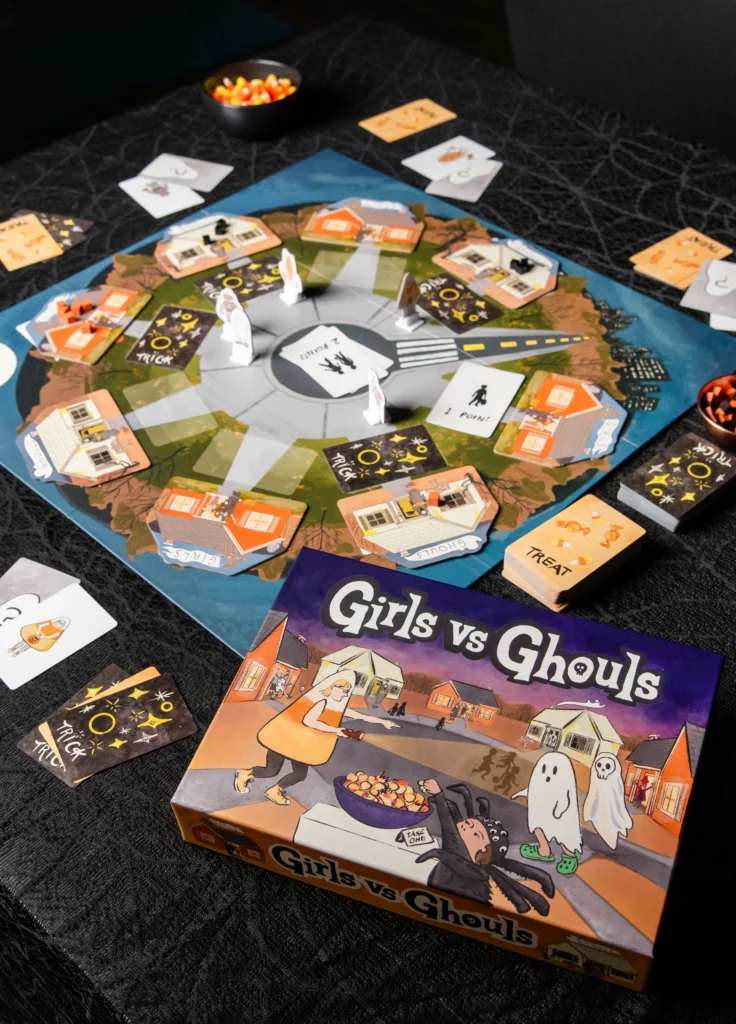 A strategy game titled "Girls vs Ghouls" is set up on a black tablecloth. The circular board displays various cards, tokens, and small ghost figures. The game box is in the foreground, showcasing a fun Halloween-themed design.